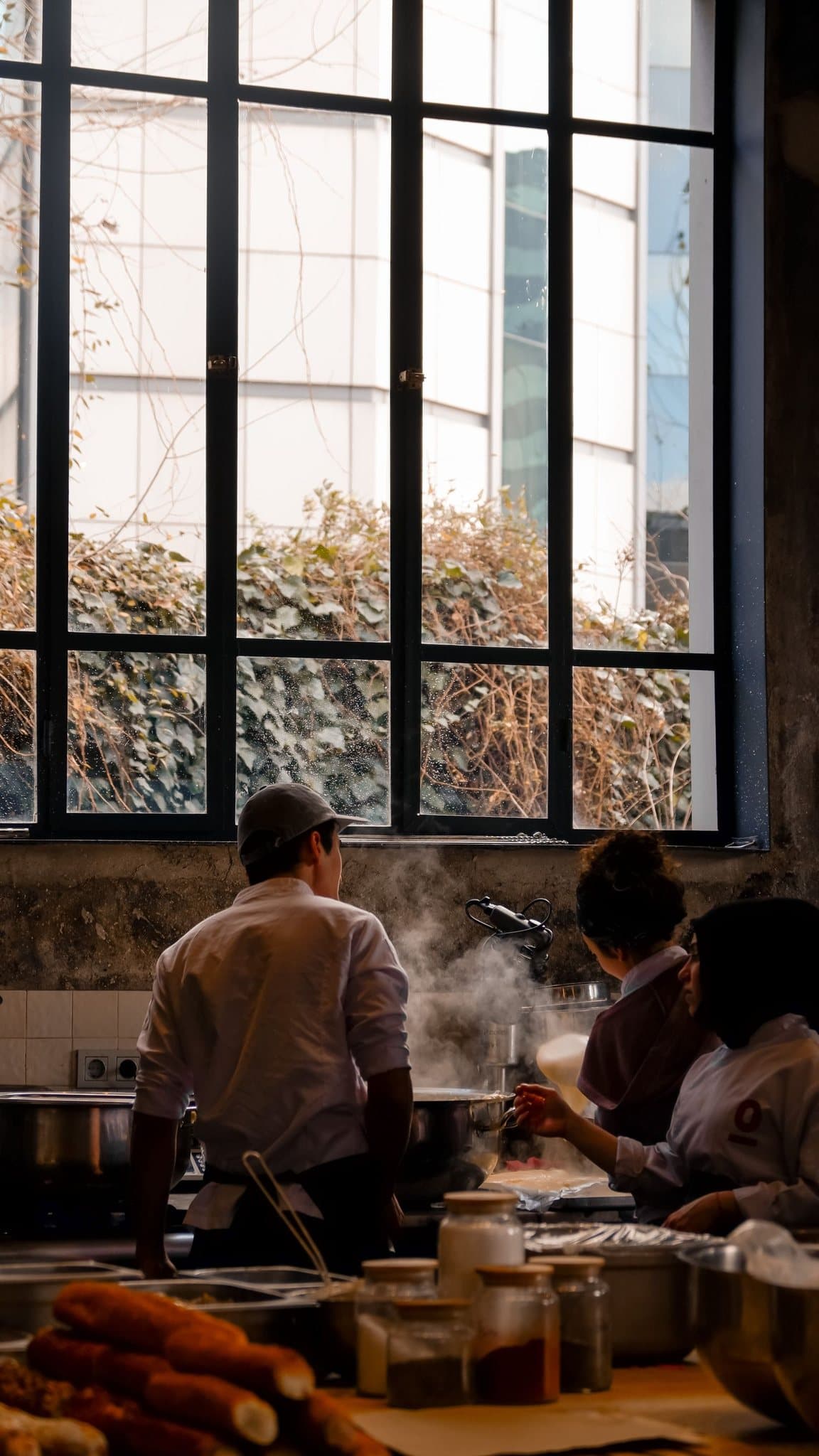 A cozinha aberta de um restaurante: cozinheiros trabalham de costas, com vapor subindo das panelas e um janelão ao fundo.